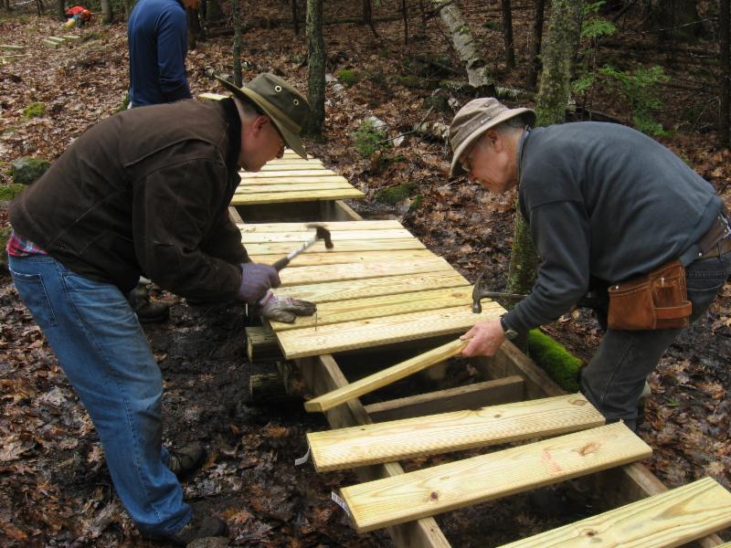 Two men banging hammers, leaning over a wooden walkway, in the forest.